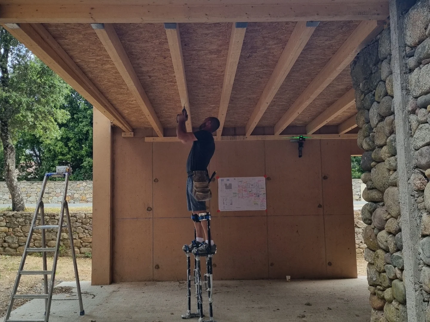 Ouvrier sur échasses fixant le plafond sous une charpente bois, plan affiché sur le mur