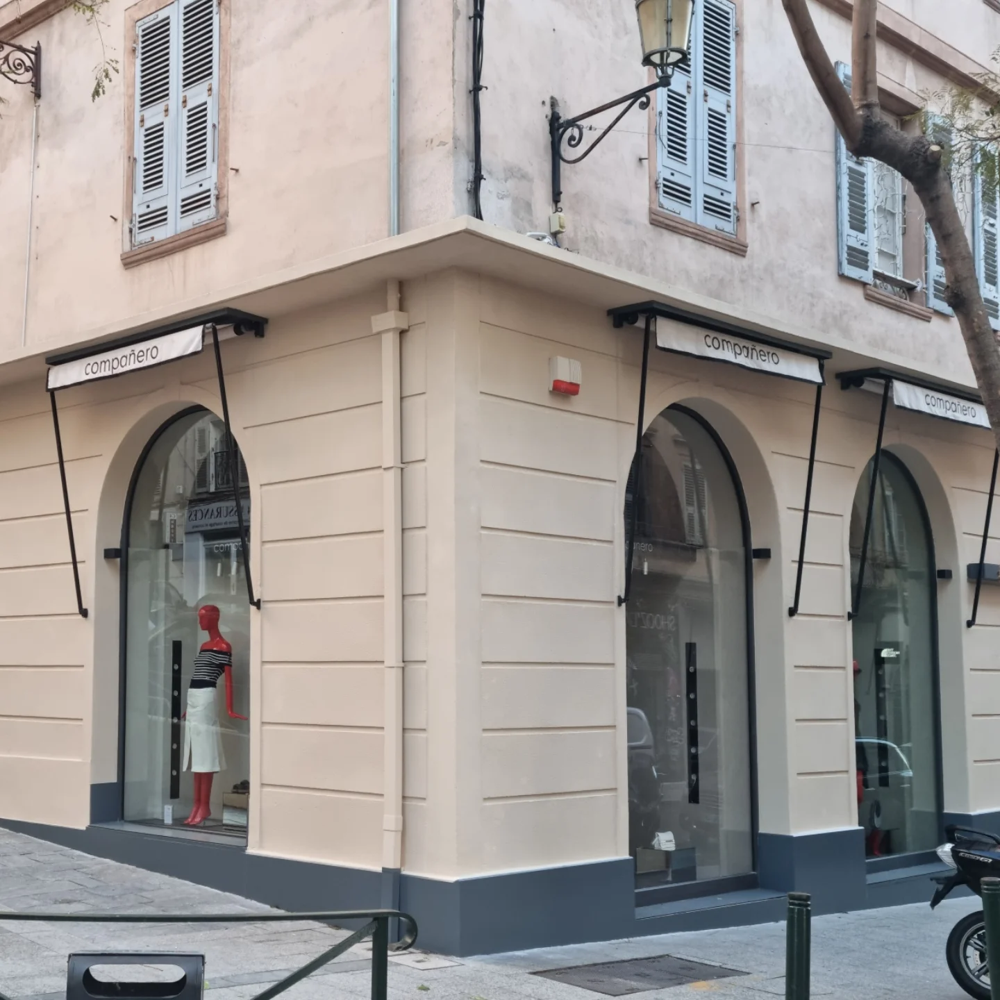 Façade d’angle d’une boutique refaite, enduit beige, soubassement gris foncé et vitrines cintrées sous des auvents
