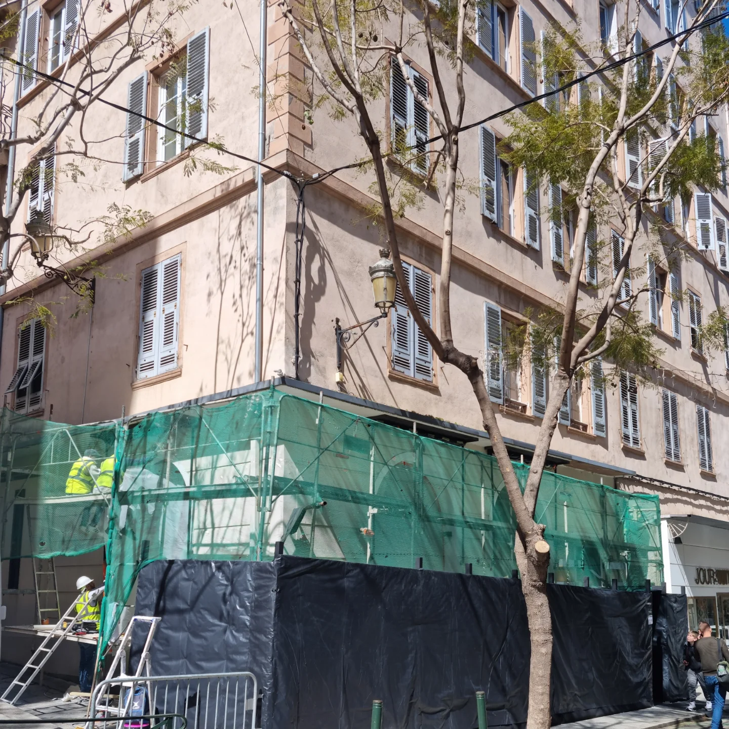 Façade de la même boutique en cours de travaux, échafaudage bâché et ouvriers au pied d’un immeuble ancien