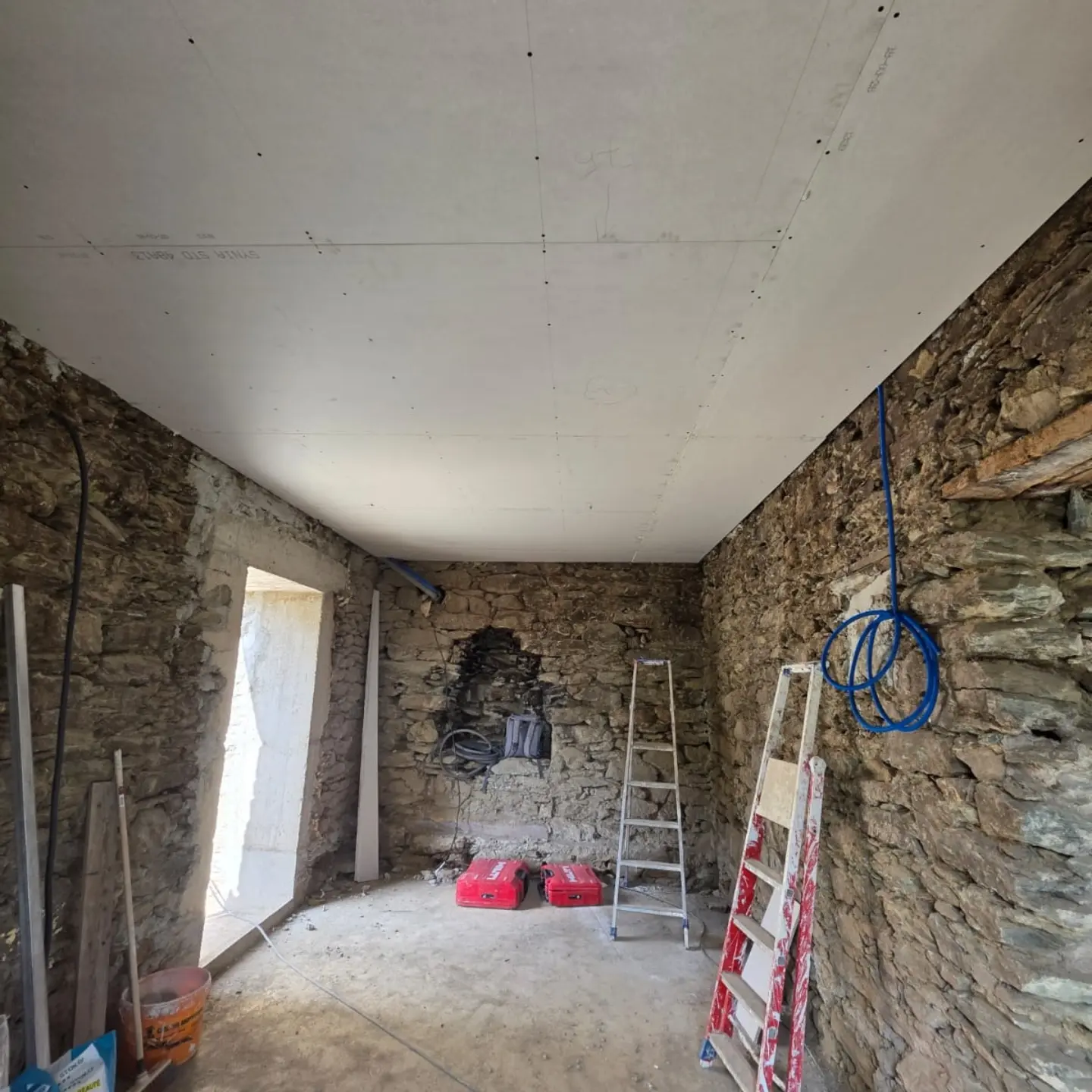 Pièce en pierre avec plafond en plaques de plâtre posé, escabeaux et outils au sol