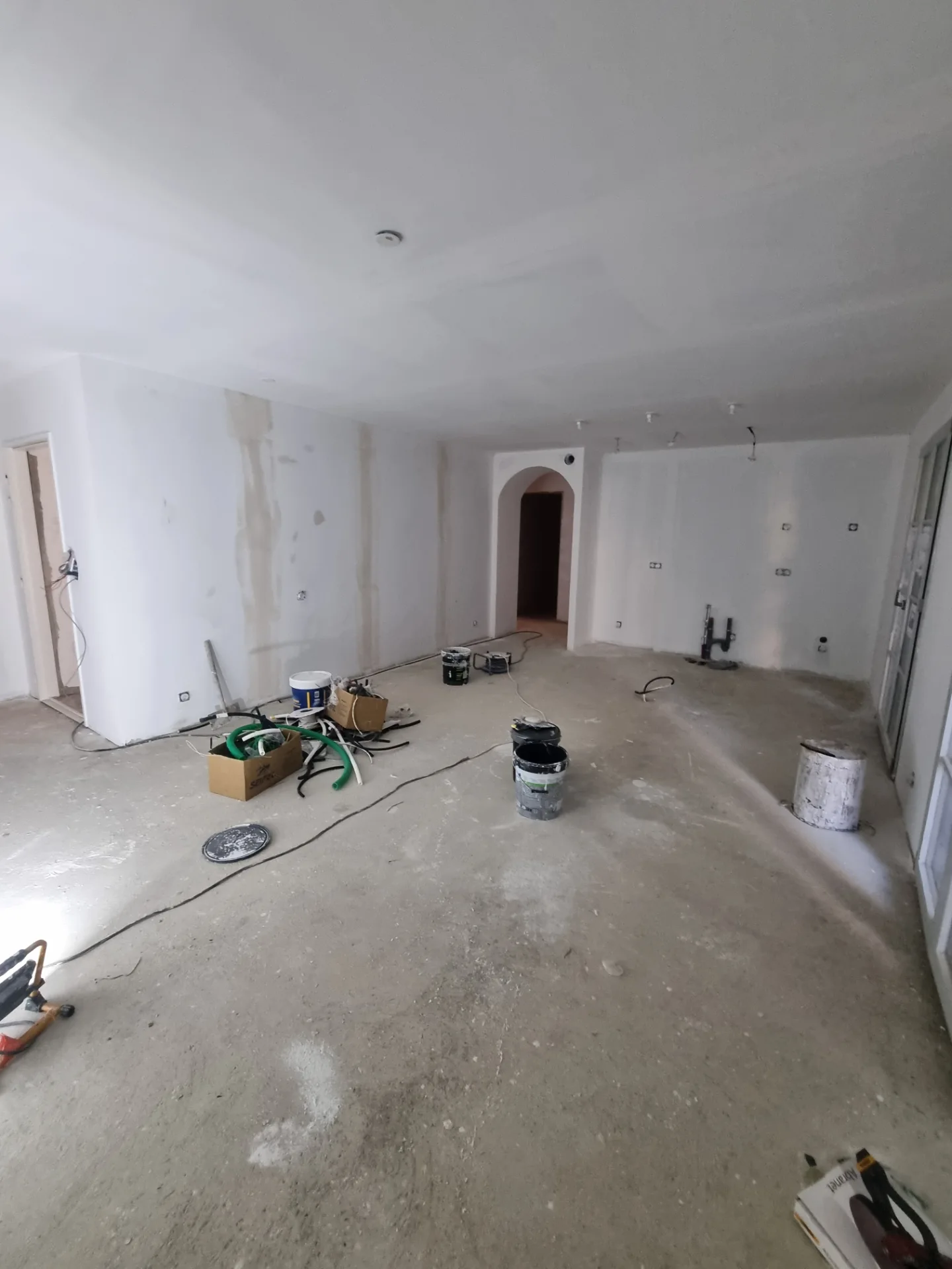 Séjour en chantier : murs et plafond enduits et blancs, arche vers un couloir, sol brut