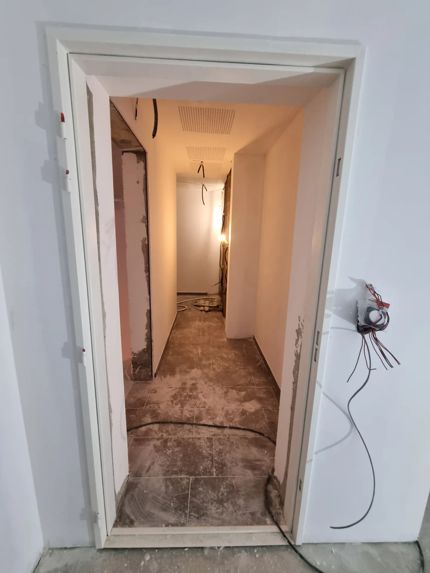 Couloir carrelé vu à travers un cadre de porte, murs enduits et plafond perforé