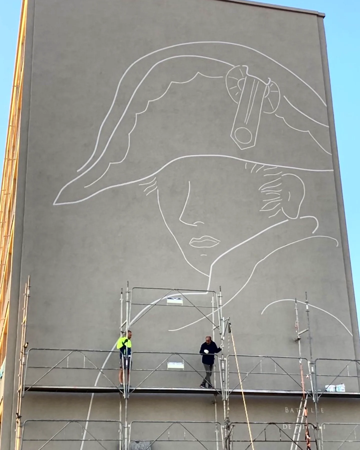 Portrait de Napoléon tracé en blanc sur le pignon gris d’un immeuble, deux ouvriers sur un échafaudage au pied du mur